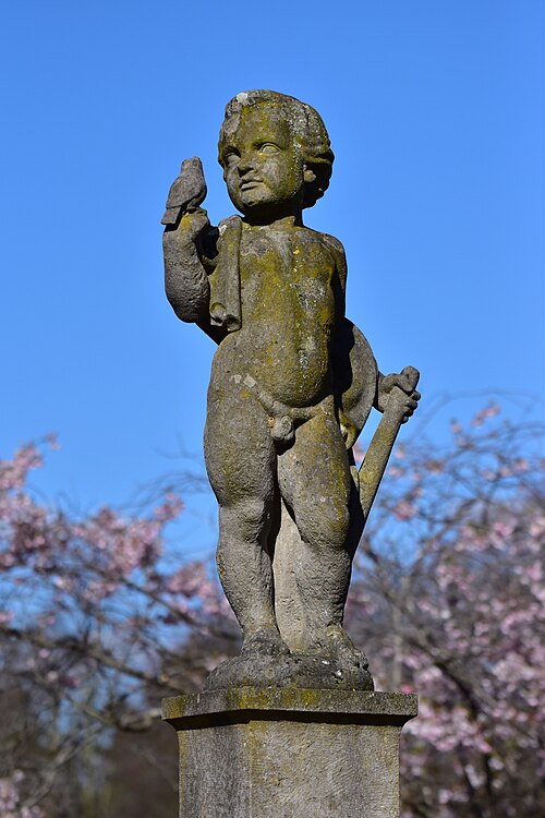 Skulptur eines kleinen Jungen mit einem Vogel auf der Hand. Im Hintergrund blühen Bäume.